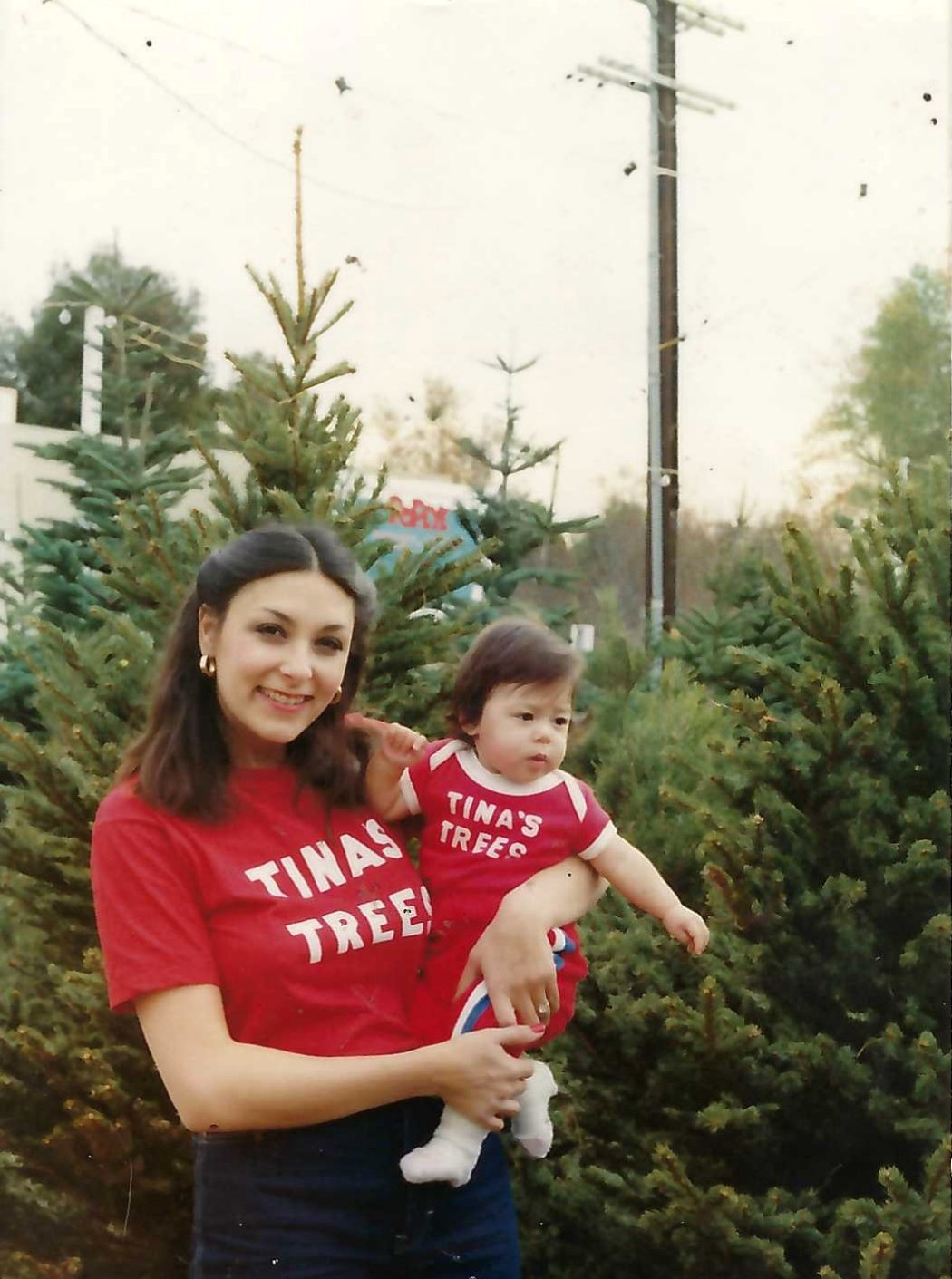 A woman holding a child, both wearing red Tina’s Trees shirts among Christmas trees